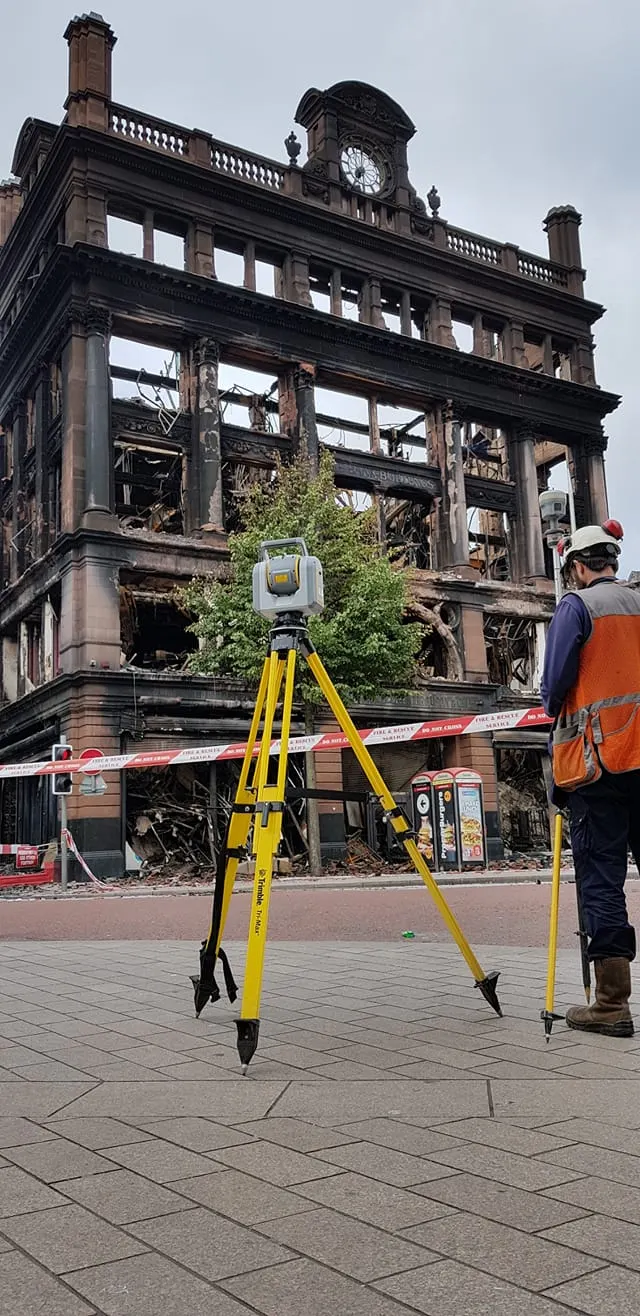 A surveyor uses equipment on a tripod in front of a fire-damaged building