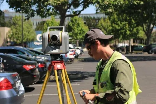 A surveyor in a high-visibility vest operates equipment on a tripod in a car park